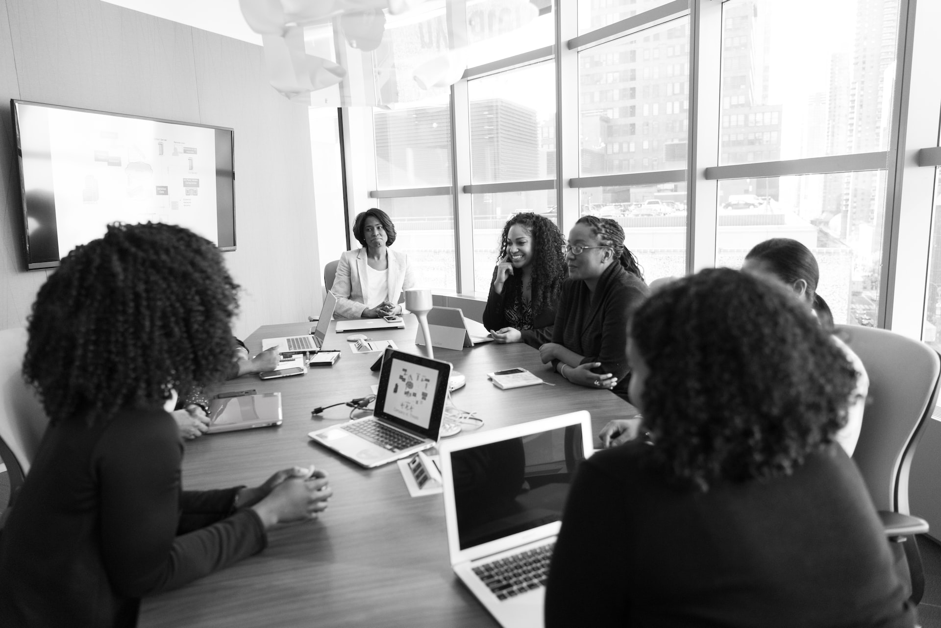 A black and white photo of women sitting around a table in a large meeting room with windows..