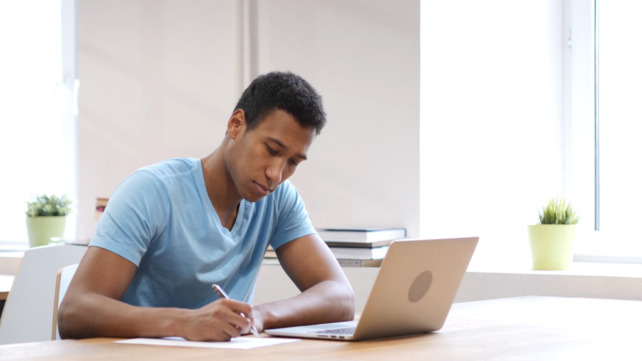 Student writing with pen at desk with laptop on desk