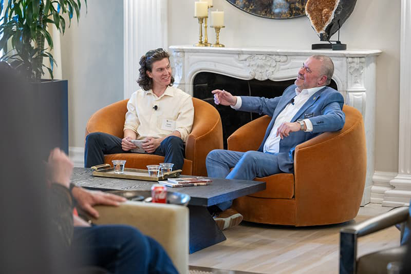 Two people seated in large brown chairs having a discussion in front of a fireplace. 