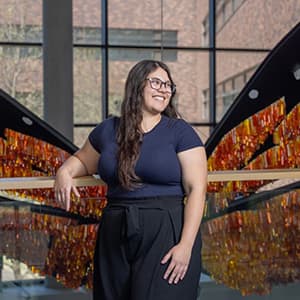 Woman standing in front of an orange and black sculpture, she wears glasses and looks off to the side as she smiles with her teeth and leans against a railing.