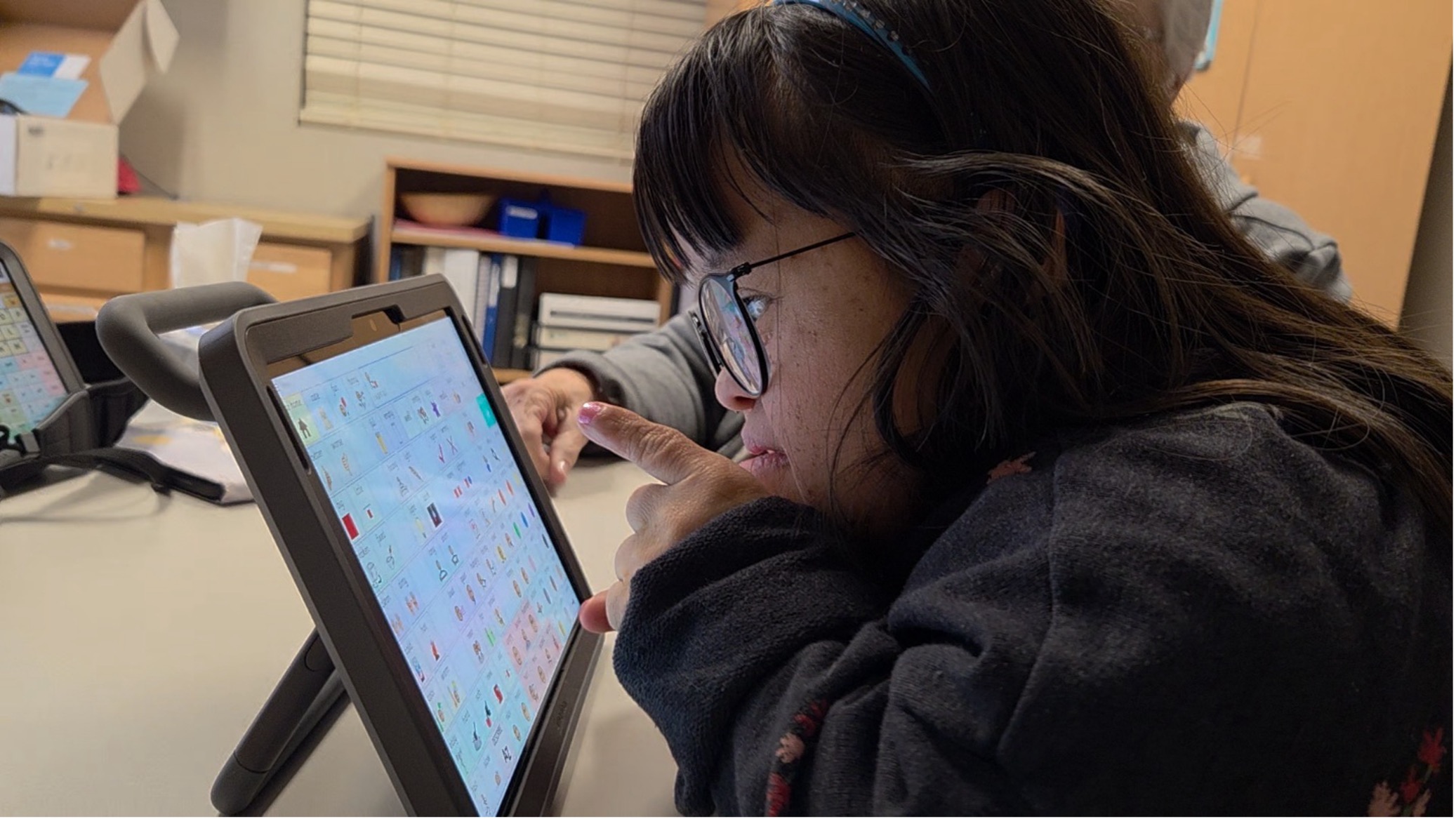 Rosie Graham uses her finger to select an icon on her AAC communication device, a tablet displaying a grid of colorful symbol icons