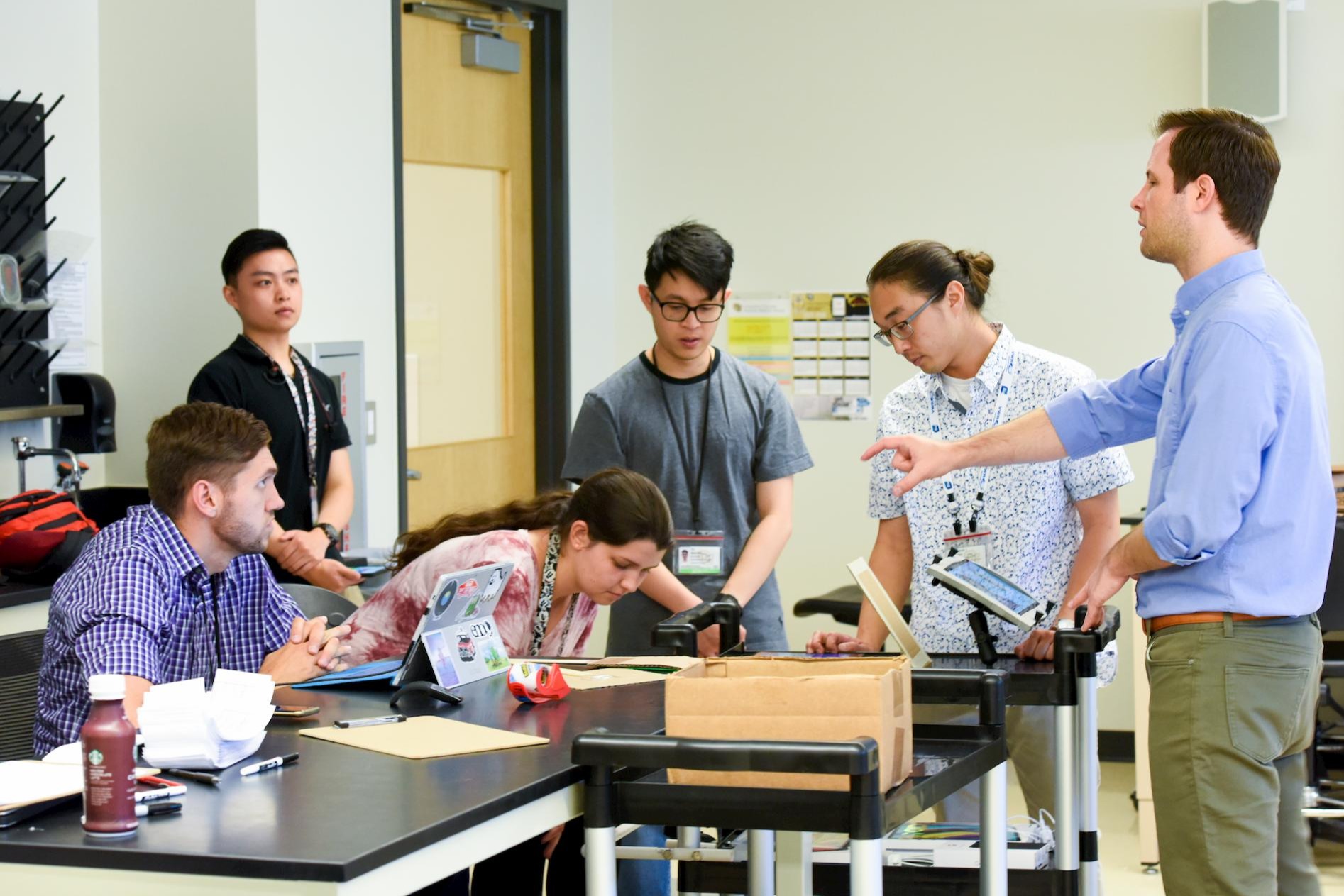 Adult students stand around a lab table talking and pointing at a box in the middle