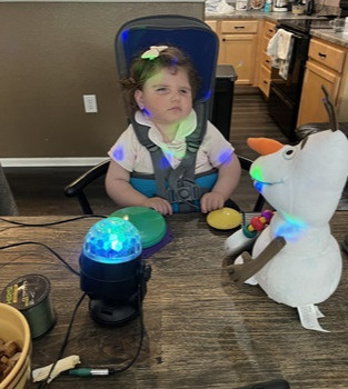 a child sitting in an adaptive seat with two switches and two toys in front of her